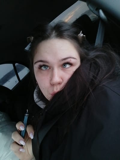 A young woman is in a car. She has dark hair with two clips in it. She's wearing a black coat pulled up around her neck, obscuring her face slightly. She holds a dark blue vape pen in her hand with light pink and white nails. She's looking to the side with a playful expression. The car window is visible in the background.