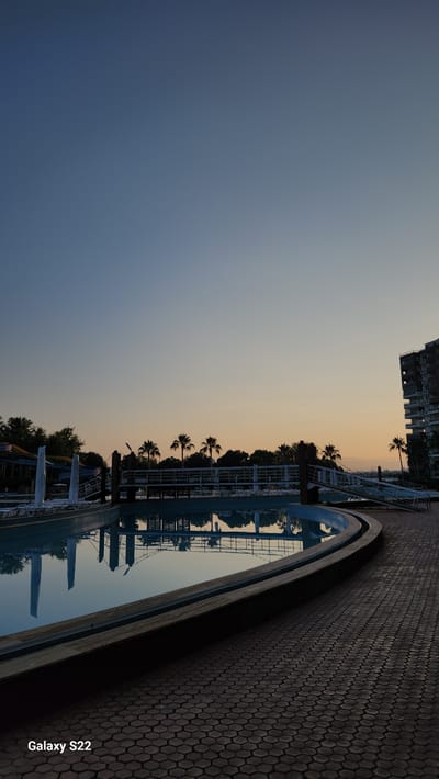 The image displays an outdoor swimming pool complex in Erdemli, Turkey, captured at either dawn or dusk. The sky transitions from a deep blue in the upper left to a pale orange-yellow hue towards the horizon on the right, indicating ambient lighting conditions. A prominent line of silhouetted palm trees stretches across the middle ground, backed by darker foliage. A dark-colored pedestrian bridge with white railings extends over a section of the pool area, reflecting in the water. To the far right, a multi-story building with multiple balconies and circular architectural features is partially visible. On the far left, colorful water slides, partially covered or wrapped, are discernible behind a section of the pool, along with several empty, white-covered lounge chairs. The main subject in the foreground is a large, irregularly shaped swimming pool with still, reflective water that mirrors the sky and surrounding structures. The immediate foreground on the right features an expanse of interlocking hexagonal dark grey pavers. No individuals are present in the scene. A "Galaxy S22" watermark is located in the bottom left corner.