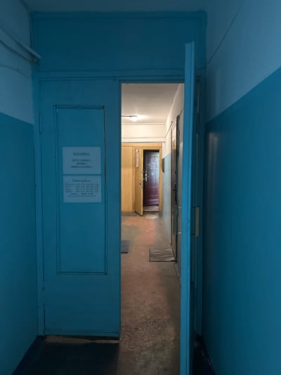 The image depicts a dimly lit interior hallway in Vilyuchinsk, Russia. On the left, a blue door is partially open, revealing a sign with Cyrillic text that reads "НОТАРИУС" (Notary) and the name "КРАСАВИНА ИРИНА НИКОЛАЕВНА" (Krasavina Irina Nikolaevna). Below the name, a "Режим работы" (Work schedule) is listed. The hallway is narrow and features blue painted walls with a white stripe. Further down the hallway, another door is visible, slightly ajar. The floor is concrete. The lighting appears to be from an overhead fluorescent fixture, casting a cool, blueish tone throughout the scene.