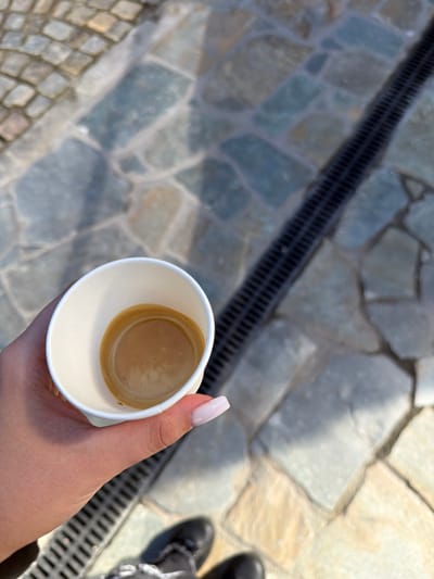 A top-down outdoor shot captures a person's hand holding a small, white paper cup filled with espresso, identifiable by its dark brown liquid and a thin layer of light brown crema. The hand features long, light pink/nude manicured nails. The background shows a textured ground surface, predominantly paved with irregular, flat, light-colored stones, possibly slate, with visible grout lines. A dark, linear drainage grate runs diagonally across the ground. In the lower-left corner, a section of what appears to be cobblestone paving is visible. A portion of a black shoe or boot, with light-colored laces or detailing, is seen in the upper-right corner. The bright, clear daylight suggests good weather, casting subtle shadows. The scene depicts a casual moment, possibly a coffee break, in an outdoor setting in Barziya, Bulgaria. No text is visible.