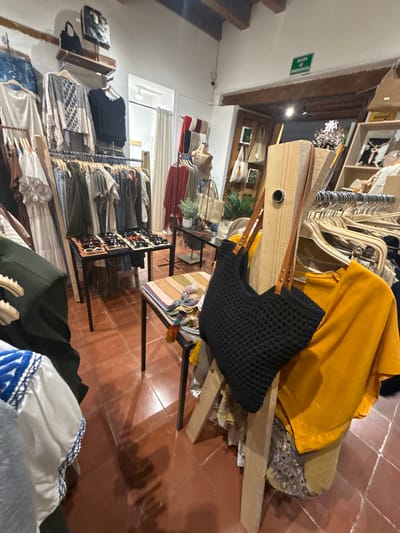 The image shows an indoor setting within a retail environment, likely a clothing or accessories store, in the city of San Cristóbal, Mexico. The floor is made of red-brown tiles. Clothing racks filled with various garments line the walls. A display table in the foreground presents sunglasses. A black crochet-style bag with brown leather straps is hanging on a wooden structure. A yellow garment is draped over a partially deconstructed chair or display. There are no people in the visible area.