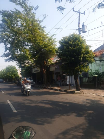 The image depicts a street scene in Pekalongan, Indonesia, on a clear, sunny day. In the foreground, a white motorcycle is being ridden by a person wearing a brown shirt and a white cap. The motorcycle's rearview mirror is visible in the bottom left corner. The road is paved with asphalt and has a white lane marking.

To the right of the road, a sidewalk is present, lined with trees. Behind the sidewalk are several buildings, some appearing to be shops or residences, with tiled roofs. A power pole with electrical transformers and wires is visible in the background. There are other vehicles, including a white SUV and a dark colored car, parked or driving on the road. Bicycles are also visible on the sidewalk. The scene suggests a typical urban environment with a mix of traffic, pedestrian activity, and overhead utility infrastructure.
