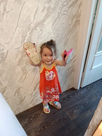 A young child, likely a toddler, is standing indoors, holding a pair of flip-flops. The child is wearing an orange sleeveless dress with a floral print and a white trim around the neckline. The child's feet are bare except for one sandal on the left foot, which is light-colored with a colorful design. In their hands, they are holding a pink flip-flop in the right hand and the sole of a sandal in the left hand. The background consists of a wall with a textured, marble-like pattern and a dark wooden floor. To the right, a white door frame is visible, suggesting the child is near an entrance or doorway. The lighting appears to be natural, coming from an unseen source. The scene is indoors, possibly a residential setting, in Shishkin Les, Russia.