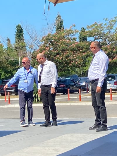 Three bald men, all wearing glasses, stand on a paved area in an outdoor location. The man on the left wears a light blue collared shirt, dark pants, and blue and white sneakers. He has a lanyard with an ID card around his neck. He is gesturing with his left hand. The man in the middle wears a white collared shirt, black pants, and black shoes. He has a dark tie. He is looking towards the man on the left. The man on the right wears a white collared shirt, black pants, and black shoes. He is looking away from the other two men. In the background, several dark-colored cars are parked. Trees with pinkish-red blossoms are visible behind the cars. The sky is clear and blue. The scene appears to be in Golubovci, Montenegro, possibly an outdoor parking area or plaza.
