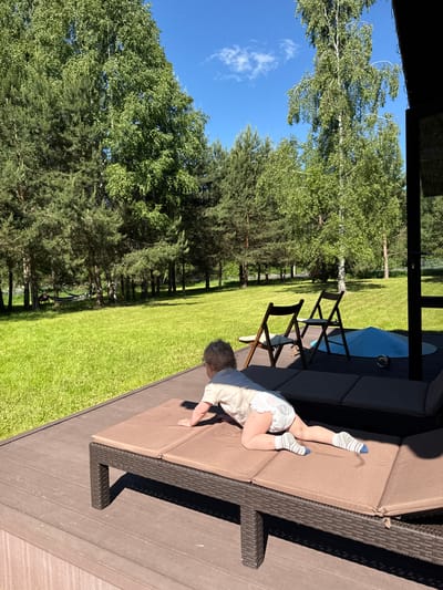 The image depicts an outdoor scene in Копнино, Russia, under bright daylight with a clear blue sky. In the foreground, a young child, appearing to be a toddler, is positioned on their stomach on a dark brown wicker-style sun lounger with light brown cushions. The child has light-colored curly hair and is wearing a light-colored short-sleeved shirt, a diaper, and white socks with blue and gray stripes. Marks are visible on the child's legs. The child's arms are extended forward, supporting the upper body, while the knees are bent, suggesting a crawling or climbing motion. The lounger is situated on a brown slatted deck or terrace, which occupies the lower portion of the frame. To the right of the lounger, two dark wood folding chairs with light cushions are visible, facing outward towards the lawn. Further to the right on the deck, a light blue conical object, possibly a recreational item, and a small metallic sphere are present. A dark structure, likely part of a building, is partially visible on the far right edge of the frame.
Beyond the deck, an expansive, well-maintained green lawn fills the midground. In the background, a dense line of various trees borders the lawn. These include numerous pine trees (conifers) on the left side and several birch trees (deciduous with light bark) scattered throughout, particularly prominent in the center-right, all with full green foliage. A dark-colored hammock is visible strung between two trees in the distant left background. The sky above is a clear blue with a few small white clouds. Strong shadows indicate a high sun position.