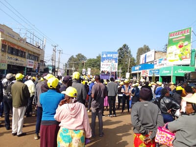 In Kimumu ward, Kenya, a large group of people gather on a paved street under a clear sky. Many individuals, predominantly women and men, are wearing bright yellow hats, suggesting a political rally or event. Some are standing in small groups, engaged in conversation, while others are facing a stage or raised platform in the background where a group of performers or speakers are visible. Various shops and businesses line the street, including "Dura Coat," "Flamingo Drycleaners," and "Safaricom," indicated by prominent signage. Billboards and large advertisements are also present, with one particularly noticeable green billboard featuring the word "REFEREE" and images of produce. Power lines and utility poles are visible overhead, and trees are interspersed with the buildings. The overall atmosphere appears to be lively and participatory, with the yellow hats creating a strong visual element amidst the crowd.