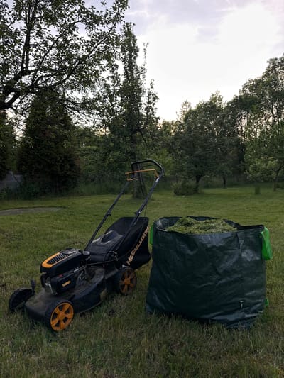 This image, captured in Staro Selo, Bulgaria, presents an outdoor scene centered on lawn maintenance equipment. A black and orange McCulloch walk-behind petrol lawnmower is positioned on mown grass in the mid-ground. The lawnmower features orange wheels and engine components, with a black grass collection bag attached to its rear. Adjacent to the lawnmower, to its right, a dark green woven plastic garden bag is filled to capacity with freshly cut green grass clippings. This bag is equipped with bright green carrying handles.
The immediate ground consists of green grass, displaying sections that have been recently mown and others that retain longer growth. The background is dominated by a dense arrangement of trees and vegetation, comprising both deciduous trees with light green foliage and darker coniferous trees. A faint wire fence is discernible through the vegetation on the left side of the frame. The sky above is overcast, characterized by light grey and white cloud formations, indicative of a diffuse ambient light condition. No human subjects are present within the depicted scene.