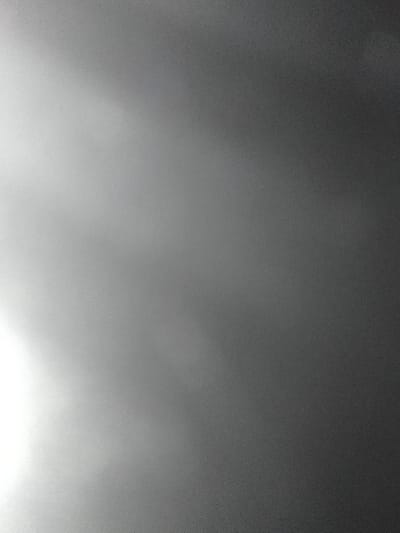 A gray gradient fills the image, transitioning from a bright white in the lower left corner to a dark, nearly black tone towards the upper right. The gradations are smooth with no defined edges. The entire composition appears slightly grainy, suggesting a photograph taken in low light conditions. The light source is implied in the lower left, creating a soft illumination across the frame.