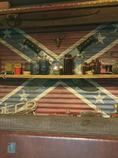 The image shows a display shelf against a background resembling the Confederate flag. The background appears to be corrugated metal painted with red and white stripes and a blue X with white stars. The shelf contains various objects, including glass bottles, a knife, a gauge, and a red plastic cup. The location is Clarksville, United States.