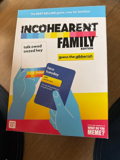 This is a close-up, eye-level, indoor photograph of the board game "Incohearent Family Edition". The game box is mostly visible, with the bottom edge of the box in focus. The game is laid flat on a wooden surface, which appears to be a floor or table. The lighting is bright and appears to be from an overhead source. The background is a wooden surface with a visible grain. The box itself features a vibrant color scheme with blue, yellow, and red sections.
The front of the box prominently displays the game title "INCOHEARENT FAMILY EDITION" in large, bold letters. Several game cards are depicted on the box. One card shows the gibberish phrase "talk owed oozed hey". Another card, partially obscured, reads "taco tuesday" with a hint below it stating "weekly meal" and a descriptive sentence "The only thing getting you through Monday". Below the main title, the phrase "guess the gibberish" is written in yellow. The bottom left corner of the box has a red rectangle with the text "AGES 12+" in white lettering. The bottom right corner features a pink section with white text that reads "From the creators of WHAT DO YOU MEME?".
The image is undated and has no discernible landmarks or features that would indicate its specific location within the city of Muskegon, United States of America. The scene is devoid of people, animals, or any other living subjects. The focus is solely on the game box and its contents as depicted on the packaging.