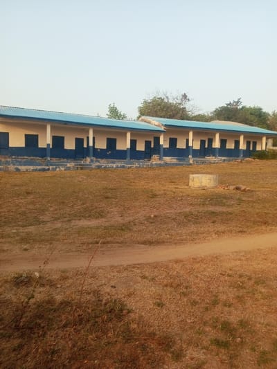 The image displays an exterior view of a long, single-story building located in Okeni, Nigeria. The building is characterized by a light blue corrugated metal roof and walls painted with an upper section of light cream or off-white and a lower section of dark blue, exhibiting visible wear and weathering. A covered veranda with supporting pillars extends along the front facade, featuring multiple dark blue doors and rectangular windows. The structure appears to be composed of at least two connected sections. The foreground and midground consist of dry, sparsely vegetated ground with patches of brown dirt and sparse grass. A dirt path curves from the lower left across the midground. A circular concrete structure, possibly a wellhead, is situated in the midground to the right of the path. Trees with varied foliage are visible in the background, behind the building. The sky is clear and light. No individuals or active subjects are present in the scene.