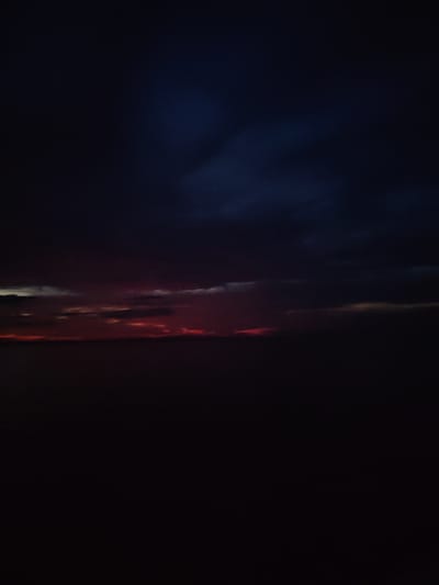 A dark, dramatic sky with hints of red and orange streaks is visible above a dark, indistinct landscape. The image appears to be a photograph of a sunset or twilight, with heavy clouds obscuring most of the light. The lower portion of the image is almost entirely black, suggesting a landscape that is either very dark or out of focus. The location is identified as Kvartsitnyi, Russia.