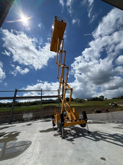 An extended yellow scissor lift is positioned on a concrete surface under a bright, sunny sky with scattered clouds. The scissor lift is partially in shadow, and its platform is extended upwards. In the background, a partially constructed building with metal beams is visible, along with a green, hilly landscape and a road. Several small excavators are present on the construction site. The scene appears to be a construction area in Janowice, Poland.