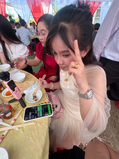 The image depicts a lively social gathering taking place outdoors in Tay Son Commune, Vietnam, beneath a festive canopy of red and white fabric. The central figure is a young woman with long, dark hair, wearing an elegant light-colored, possibly cream or white, flowing dress. She is smiling and making a peace sign with her right hand, displaying a silver watch, gold rings, and a delicate gold necklace, conveying a cheerful and relaxed mood.
In front of her, on a table covered with a yellow cloth, a white smartphone is prominently displayed. Its screen shows an application with various details: "Ruby," "RAPPER," "Your mapper rank is #1801," "13.5K," "0," "$0," "$0," "$29.84," "$10.89," "Get 20 PATTN," "Collect your Creator's Reve," "Withdraw ATTN," "Swap," and "Successfully collected 90.18 PRTTH from I nenories." Adjacent to the phone are several white bowls, some containing food remnants, along with wooden chopsticks, a pink selfie stick or tripod, and a clear glass decorated with colorful polka dots. A small pink handbag also rests on the table.
To the left, another woman in a red outfit is partially visible, actively eating with chopsticks. In the blurred background, numerous other individuals, primarily men in light-colored shirts, are seated at additional tables, participating in the event. The scene suggests a casual daytime meal or celebration, characterized by communal dining and friendly interaction.
