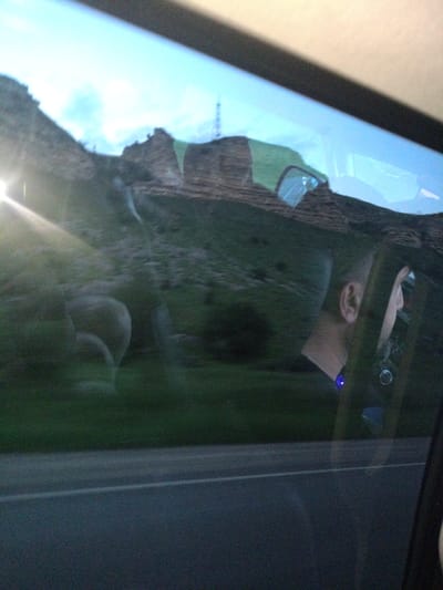A person's head and shoulder are visible in the reflection of a car window. The person has short dark hair and is wearing a blue device on their neck. The reflection also shows a landscape of rolling hills with rocky outcrops and a bright light source, possibly the sun, in the upper left corner. The car is on a road, suggested by the blurry asphalt in the foreground. The scenery appears to be mountainous. Given the context provided, the location is estimated to be near Ali-Berdukovskiy, Russia, a settlement known for its mountainous terrain.