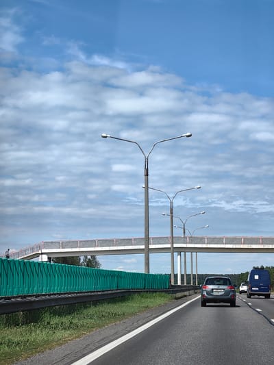The image, captured in Негарэльскі сельскі Савет, Belarus, depicts a multi-lane asphalt highway scene during daylight hours. The upper portion of the frame shows a blue sky with scattered white cumulus clouds.
In the foreground, the highway has at least two visible lanes in the direction of travel, marked by a dashed white line. A solid white line defines the left edge of the paved shoulder. A dark gray Subaru Outback station wagon, bearing Belarusian license plate "4955 TM-7," occupies the rightmost visible lane, traveling away from the viewer. Further down the road in the same lane, a blue Mercedes-Benz Sprinter van is visible, followed by a partially obscured white car.
Along the left side of the highway, a metal guardrail runs parallel to the road. Behind the guardrail is a tall green sound barrier constructed from numerous vertical panels. A strip of green grass grows between the guardrail and the road's edge.
Several streetlights with concrete posts and silver-colored, bifurcated arms extending over the highway are positioned along the left side.
A concrete pedestrian overpass spans horizontally across the highway. The overpass features light-colored railings with red top bars and rectangular grated infill panels. It is supported by multiple concrete pillars. On the far left side of the overpass, a lone individual, dressed in dark attire, is visible ascending a ramp leading to the overpass structure.
The background beyond the overpass reveals a distant line of dense green trees, indicating a forested area.