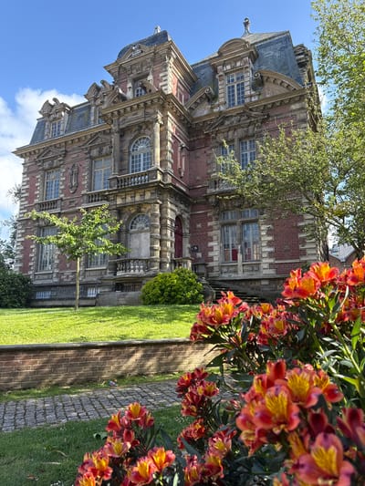 A vibrant outdoor scene features a grand, multi-story building alongside a lush garden under a clear blue sky in Fécamp, France. The building, prominent in the left and center, exhibits intricate architecture with red brick walls, grey stone details, ornate carvings, balustrades, and dark slate roofs. Numerous windows of varying shapes, including arched and rectangular, are set within decorative frames. Several large trees with bright green foliage partially obscure the upper sections of the building.
In the foreground on the right, a profusion of bright orange and yellow flowers, likely Alstroemeria (Peruvian Lilies), adds a striking splash of color and depth to the composition. Below the flowers, a well-maintained green lawn stretches out, bordered by a rustic brick path. Adjacent to the brick path, another path made of grey cobblestones runs parallel, creating a clear demarcation within the garden space. A small, slender tree with light green leaves stands in the grass closer to the building.
The scene is bathed in bright sunlight, indicated by the crisp shadows cast on the ground and building, suggesting a clear and sunny daytime. The sky is a vivid blue with a few scattered white clouds. No specific activity is depicted, presenting a static, picturesque view of the historic architecture and natural beauty. No legible text is visible in the image.
