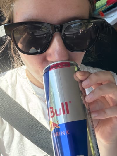 A close-up, eye-level, vertical shot shows a person wearing black sunglasses and a white t-shirt, with a seatbelt across their chest, drinking from a Red Bull can. The person's face is partially obscured by the can and sunglasses. The sunglasses reflect a blurred image of a car interior and trees. The Red Bull can is silver with red and blue lettering, and the Red Bull logo is visible. The person's right hand, with clean fingernails, is holding the can. The background is out of focus, showing what appears to be a car interior and possibly an outdoor scene through a window. The scene takes place inside a vehicle, likely on a road. There is no indication of the city of Seaside, United States.