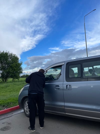 A man in a dark-colored hoodie and pants is standing next to a silver van, leaning forward and looking into the driver's side window. The van is parked on the side of a road next to a grassy area with trees. The sky is blue with scattered clouds. A tall light pole is visible in the background. The location is near Бавыкино, Russia.