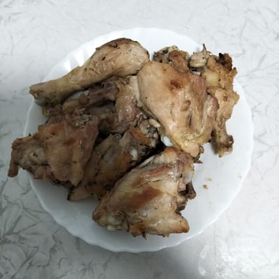 A close-up, overhead shot displays a white, scalloped plate filled with an assortment of cooked chicken pieces. The chicken appears to be seasoned with black pepper and has a slightly browned, glossy appearance, suggesting it has been roasted or pan-fried. The pieces vary in size and cut, including what look like drumsticks, thigh portions, and possibly wing sections. Some pieces show exposed bone. The plate rests on a white tablecloth with a subtle, textured pattern resembling marbled paper. The lighting is even, with no strong shadows, and the background is out of focus. The image is a still life of food, with no indication of its location in Cupcini, Moldova.