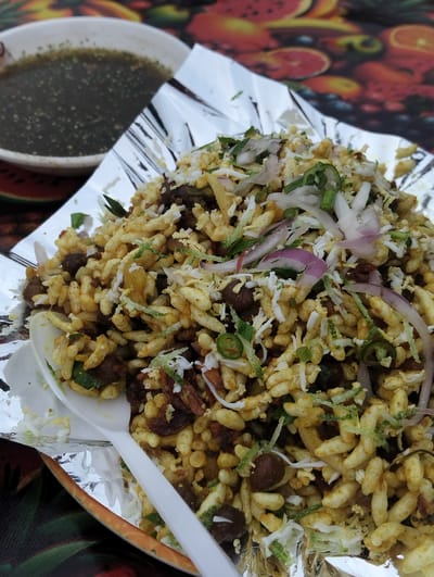 A close-up, eye-level shot shows a serving of Jhalmuri, a popular Bengali street food, placed on a disposable aluminum foil lined plate. The Jhalmuri is a mixture of puffed rice, chickpeas, chopped onions, cilantro, and spices, topped with shredded white cheese and thinly sliced green chilies and red onions. A white plastic spoon rests on the left side of the plate, partially submerged in the Jhalmuri. In the upper left quadrant of the image, a white bowl contains a dark, liquid-based mixture, possibly a chutney or sauce, with visible green specks. The background is out of focus, revealing a colorful, patterned surface that appears to be a tablecloth, with abstract shapes resembling fruit slices. The lighting suggests an outdoor setting, possibly a street food stall in Purbachal, Bangladesh.