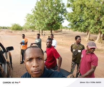 The image is a selfie taken in Gidan Kogi, Nigeria, featuring seven men of African descent. The selfie taker is in the foreground, wearing a navy blue shirt, with a serious expression. Behind him, a man in a red t-shirt sits on a motorcycle. Further back, there are five other men wearing various shirts, including orange, green, and white. A black car is partially visible on the left side. The background includes a dirt road and trees. In the distance, there are dwellings and other people visible. The picture was taken with a Camon 40 Pro, 20mm f/2.4 1/139s ISO151.