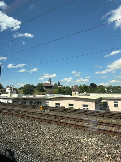 The image captures a sunlit landscape from what appears to be a train window, looking towards a semi-urban and elevated area under a bright blue sky with scattered cumulus clouds. Multiple black overhead catenary lines stretch horizontally across the upper third of the frame.
In the foreground, train tracks filled with dark grey ballast dominate the lower half of the image. At least three distinct tracks are visible, running parallel to each other. A small yellow marker post with a circular top is positioned between two tracks on the right side. On the far bottom left, a section of a metal mesh fence is partially visible.
Immediately behind the tracks, a row of light-colored, single-story industrial or commercial buildings with flat roofs extends across the midground. These structures have numerous rectangular windows and pale facades, one of which appears slightly pinkish. One building on the right features a small, ornate, brown-roofed tower with a pointed top, distinct from the main structure.
Further in the background, on an elevated green hill, several residential houses are interspersed among dense trees. A prominent brown-roofed building with a tall, pointed spire and a clock face stands atop the hill, resembling the Reformed Church of Wetzikon. This church-like structure is situated amidst trees, with what appears to be a large barn-like building adjacent to it on the left. The scene is devoid of people.
The specific church with its distinctive spire and hillside location, viewed from the railway, strongly indicates the scene is located in Wetzikon (ZH), Switzerland, likely from a train passing near the Wetzikon railway station looking towards the Kirchhügel (Church Hill) where the Reformed Church of Wetzikon is situated. The time of day is clear and sunny, indicative of midday or early afternoon.