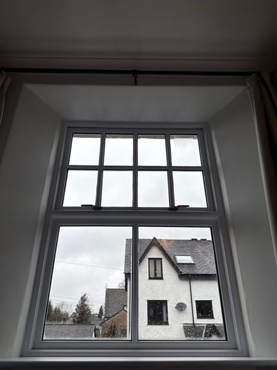 The image shows a window view of a building exterior. The window is divided into multiple panes. The exterior building is white with a dark roof. There are windows and a satellite dish visible on the building. Raindrops are on the window. The location is likely Windermere, United Kingdom. There are no people visible in the image.