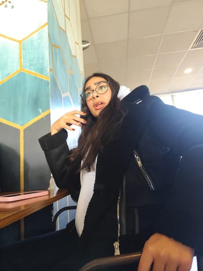 A young woman with long dark hair and glasses sits indoors. She wears a black jacket over a white hooded sweatshirt. Her left hand is raised near her face, and she appears to be looking upwards with a slightly open mouth. A wooden table with a pink cell phone on it is to her left. A geometric blue and gold design is painted on a pillar behind her. The ceiling is visible, showing white tiles and an air vent.