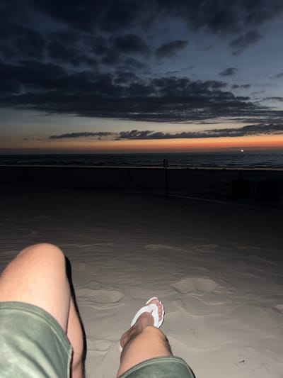 The image depicts a low-angle, personal perspective from a person sitting on a sandy beach at dusk in Langeoog, Germany. The foreground is dominated by the person's legs, clad in olive green shorts, and one foot wearing a white flip-flop, resting on the sand. The sand itself shows subtle undulations and footprints, indicative of a natural beach environment.
The middle ground of the image is a dark expanse of the beach, stretching towards the sea. Several dark, silhouetted beach chairs or huts are faintly visible in the distance, suggesting a public beach area. A thin, dark line, likely a path or boardwalk, runs across the beach.
The background is dominated by the ocean and the sky. The sea is a dark, calm expanse, with a subtle line of foam marking the edge of the water. Above the sea, the sky transitions from a vibrant orange and yellow band at the horizon, characteristic of sunset or twilight, to darker blues and grays higher up. Dark, textured clouds are scattered across the upper portion of the sky. A single, bright star or planet is visible in the sky above the horizon line. The overall impression is one of serene contemplation in a natural setting during the transitional light of dusk.
