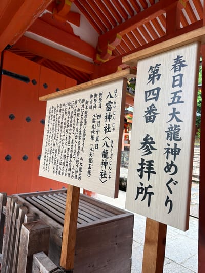 The image displays a detailed view of a worship area at the Hachirai Shrine (八雷神社), which is part of the Kasuga Taisha Shrine complex in Nara, Japan. Two prominent wooden signs with black Japanese characters stand in the foreground. The larger sign on the right identifies the location as the "Fourth Worship Place" (第四番 参拝所) for the "Kasuga Five Great Dragon Gods Pilgrimage" (春日五大龍神めぐり). The smaller sign on the left provides information about the Hachirai Shrine, identifying the enshrined deity as the "Eight Dragon God" (八龍神様), with smaller text detailing its origin and significance, including the regular festival date of April 15th. Behind the signs, vibrant orange-red traditional Japanese architectural elements, including structural beams and intricate ceiling components, are visible. In the lower left, a wooden offering box with horizontal slats on its lid is partially visible behind a low wooden railing, resting on a light grey, paved surface. The scene is devoid of people.