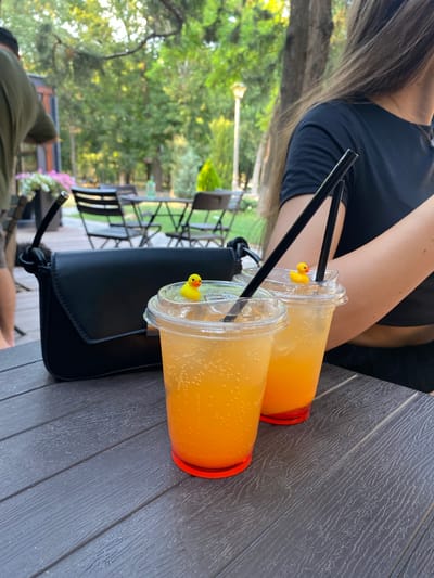 This image captures an outdoor scene at a cafe with two drinks. The subjects are a person, whose shoulder and arm are visible, wearing a black top, and two clear plastic cups filled with an orange beverage. The drinks have black straws and small yellow rubber ducks clipped to the rims, adding a playful detail. A black purse is placed in front of the drinks.

The setting appears to be a cafe with wooden tables and chairs visible in the background, along with lush green trees and foliage, suggesting a pleasant outdoor environment. The lighting indicates it is daytime, possibly late afternoon given the warm glow on the drinks. The overall mood seems relaxed and leisurely. There is no visible text in the image. The presence of drinks and the outdoor seating arrangement suggest a casual outing or a break at a cafe.
