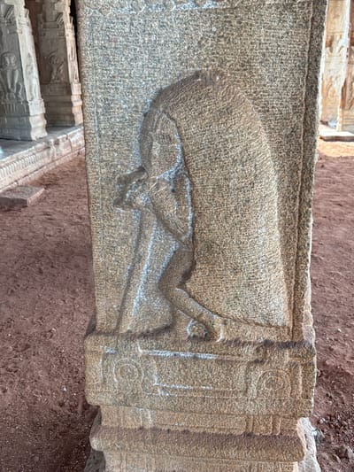 A close-up, upward-tilted shot captures a section of a weathered stone structure featuring a bas-relief carving, presumably located in or near Kamalapura, India. The carving depicts a horizontal, stylized figure, possibly an animal or mythical creature, sculpted into the stone's surface. The subject's posture suggests a reclining or falling position. The stone is textured with a coarse, grainy surface, indicating its age and exposure to the elements. To the right of the carving, the stone is fashioned into decorative columns or pilasters with carved linear and geometric motifs. The background is predominantly reddish-brown soil.