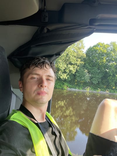 The image shows a close-up, rotated selfie of a man wearing a bright yellow safety vest over a black t-shirt. He has wet, dark hair and a light stubble. His eyes are open, and he appears to be looking directly at the camera with a neutral expression. The background is out of focus, but it suggests an outdoor setting with green trees and a body of water that reflects the trees and sky. A black curtain or fabric is visible in the foreground, partially obscuring a car's interior. The lighting suggests it is daytime, likely sunny, given the bright reflection on the water and the man's illuminated face. The location context provided is Edewecht, Germany, which might imply the setting is a canal or river typical of the region. There is no visible text in the image. The man's attire, particularly the safety vest, hints at a possible work-related activity or a recreational activity where safety gear is required, such as boating or outdoor labor. The overall mood is casual and personal, typical of a selfie.