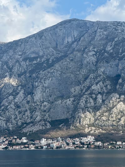 The image depicts a panoramic view of a coastal settlement in Bogdašići, Montenegro, positioned at the base of a large, rugged mountain range fronting a body of water.
The upper portion of the frame features a partially cloudy, light blue sky with white cumulus cloud formations. The dominant feature is a massive, steep, grey-blue limestone mountain occupying the middle and upper-middle ground. This mountain displays a highly fractured and eroded surface with numerous rock outcrops, vertical striations, and shadowed crevices. Sparse, low-lying vegetation, appearing dormant or evergreen, dots the mountain's slopes. A distinct, horizontally elongated, oval-shaped cave opening or erosion feature is visible on the upper-left flank of the mountain face. Towards the lower slopes, denser patches of vegetation are present, and at least two power line poles are discernible.
At the immediate base of the mountain, a dense cluster of light-colored buildings forms a town along the narrow coastal strip. These structures are predominantly white or cream-colored, with roofs of various hues including reddish-brown and grey. The buildings appear multi-storied and are tightly packed along the waterfront, extending slightly up the initial incline of the mountain.
The foreground is occupied by a calm, dark blue-grey body of water, indicative of a bay or fjord, which directly abuts the built-up area of the town. The water's surface shows minor textural variations from ambient reflections. The scene is illuminated by mixed lighting conditions, with sunlight highlighting specific sections of the mountain and town while other areas remain in shadow. No individuals or vehicles are discernable.