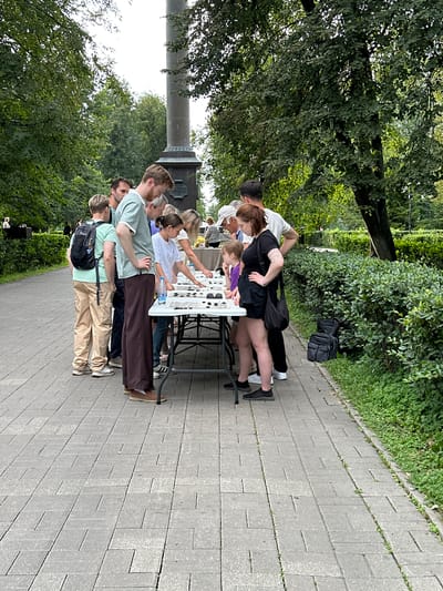 The image shows a group of people gathered around a table outdoors, possibly in a park. The table is covered with small items, and several individuals appear to be examining them. The background features trees, a tall monument or column, and a paved walkway. The people are casually dressed. There are no visible signs or indications that this scene is located in the city of Putyatino (Semivragovskaya S/A), Russia.