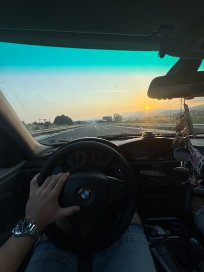 The image depicts a first-person perspective from inside a moving vehicle, specifically a BMW, traveling on a multi-lane highway at sunset in the vicinity of Ignatievo, Bulgaria.
The driver's right hand is positioned on the black, three-spoke steering wheel, which prominently features the blue and white BMW logo at its center. A metallic watch with a dark face is visible on the driver's left wrist. Below the steering wheel, the driver's legs are visible, clad in blue denim jeans.
The car's interior features a dark dashboard, with visible instrument gauges including a speedometer marked up to 260 km/h and a tachometer. The center console below the dashboard contains climate controls, an entertainment unit, and a small display screen. Keys are present in an open compartment near the gear shifter. A mobile phone appears to be placed in a holder on the dashboard.
The windshield is partially obscured by numerous small spots or smudges, indicating dirt. The upper portion of the windshield exhibits a distinct teal-colored tint. A rearview mirror is centrally positioned, with multiple pairs of sunglasses and various decorative charms hanging from it.
Beyond the windshield, the highway extends into the distance under a clear, warm-toned sky. The sun, a bright yellow orb, is low on the horizon, slightly to the right of the center frame, indicating sunset. The landscape consists of a highway with guardrails on both sides, bordered by green vegetation including trees and shrubs. In the background, distant mountains or hills are visible. Utility poles and power lines traverse the scene on the right side. An empty billboard frame is situated on the left side of the highway, while another large sign structure is dimly visible further down the road on the right.