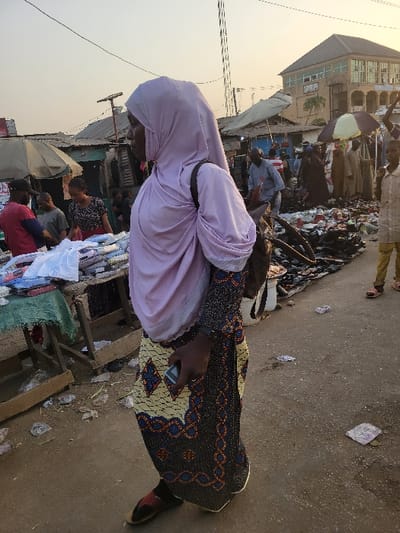 A woman wearing a light pink hijab and patterned black and yellow dress stands in a bustling marketplace. Her hand holds a cell phone. Stalls with various goods line the scene, crowded with people. A building with multiple windows is visible in the background, partly obscured by a market umbrella. Overhead power lines crisscross the sky, adding to the busy atmosphere. The ground is littered with debris.