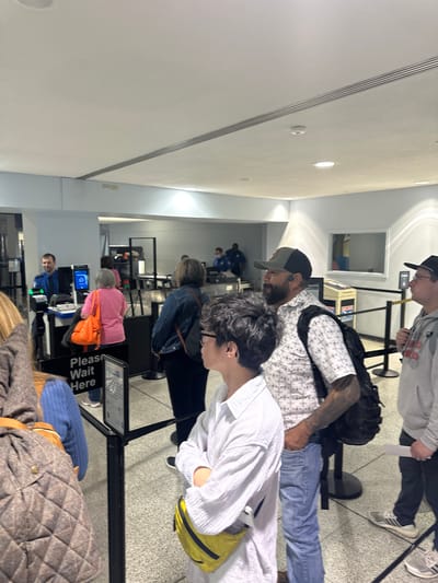 The image depicts a line of people waiting at a security checkpoint, likely in an airport or transportation hub. Individuals are standing behind retractable belt barriers with "Please Wait Here" signs. At least two security personnel are visible in the background, one operating a computer terminal and another overseeing the line. Passengers are dressed in casual attire, with some wearing backpacks or carrying bags. The environment suggests a modern, indoor space with fluorescent lighting and tiled flooring. There's no explicit confirmation of the location as Cleveland, Ohio, but based on the context, it is plausible.