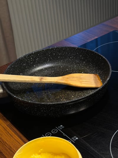 This media file shows a close-up, overhead view of a black frying pan with a speckled pattern, containing a wooden spatula. The pan is placed on a black glass ceramic stovetop, which has white markings indicating burner controls. To the right of the pan, a yellow bowl filled with a bright yellow substance, possibly butter or ghee, is visible. The scene is set in a kitchen, with a wooden countertop and a portion of what appears to be a blinds-covered window in the background. The lighting suggests it could be daytime, with natural light illuminating the scene. No people are visible, and the overall impression is one of preparation for cooking. The location is identified as Słupsk, Poland.