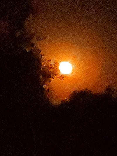 The image displays a bright, luminous sun, predominantly white with a warm orange glow, partially obscured by dark, textured clouds. The overall scene is dark, with the sun being the primary light source. The clouds have a rough, almost fuzzy texture and are cast in shades of dark brown, orange, and black, creating a dramatic contrast with the sun's brilliance. The background is uniformly dark, suggesting it is either nighttime or a very overcast day. The image appears to be a photograph of the sun setting or rising behind clouds, creating a striking visual effect. No specific location in the city of Valverde de Leganés, Spain, can be identified from the provided image.