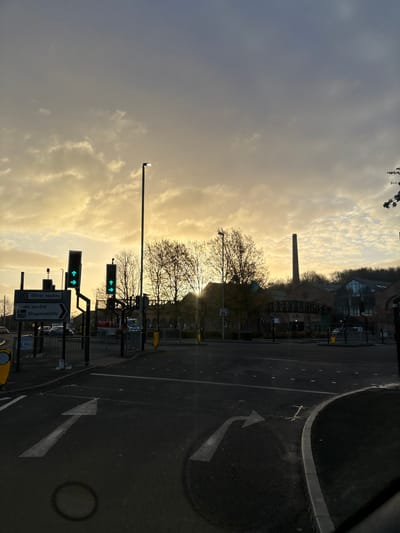 The image depicts a street scene in Oswestry, United Kingdom. The view is of a road intersection at sunset or sunrise, with the sky filled with dramatic, golden-tinged clouds. A tall brick chimney stands prominent in the background, likely part of a former industrial complex, alongside more modern structures. Traffic lights display green signals. Road markings include directional arrows. Trees with bare branches line the street. Signage indicates directions to the town center and facilities for disabled people. A lamp post stands tall to the left.