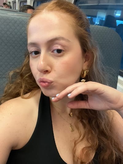 A young adult female with reddish-brown wavy hair and fair skin is depicted in a close-up, selfie-style photograph. Her lips are pursed, and her right hand, with light blue manicured nails and a gold-toned ring on the ring finger, is resting under her chin. She wears a black halter-neck top. Accessories include a gold-toned hoop earring with a dangling charm in her right ear, and two thin gold-toned necklaces; one features an infinity symbol, and the other has a rectangular pendant with the letter 'E' and a small butterfly charm. The background shows the interior of a passenger train car with dark gray patterned seats and windows. Two indistinct individuals are partially visible in the blurred background. The scene is located in a train within Secaucus, United States.
