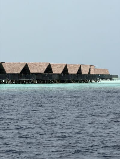 The image depicts a building in Guraidhoo, Maldives, positioned vertically. The building features a series of stepped or tiered sections constructed of a rough, light brown material. A scaffolding structure runs along the side of the building. Adjacent to the building is an expanse of water, possibly the ocean, with a textured surface. The sky is a pale, uniform blue.