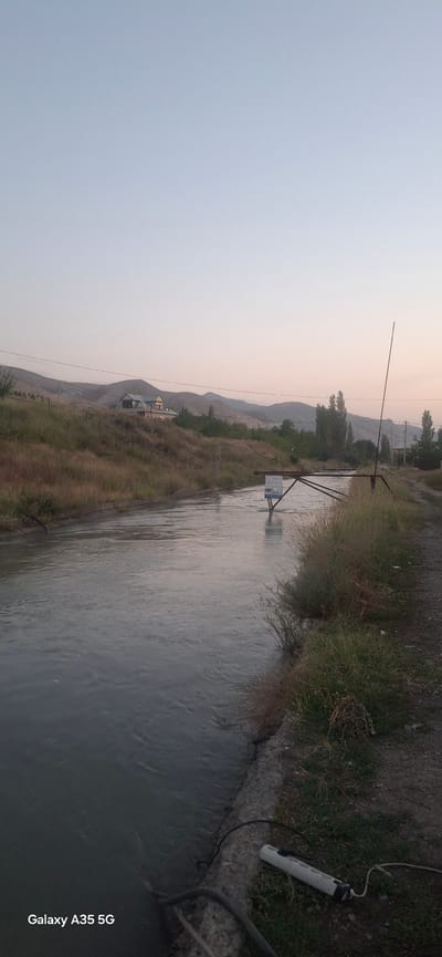 The image depicts a tranquil canal flowing through a rural area, possibly in or near Djiyde, Kyrgyzstan. The water is calm, reflecting the soft, pale sky of what appears to be early morning or late evening. On the left bank of the canal, dry grasses and a gentle slope lead up to a house with a distinct roofline. In the distance, rolling hills form the backdrop. The right bank of the canal features a dirt path, overgrown with grass and weeds, and a metal structure that spans the water, possibly a bridge or a control gate. A tall, thin pole stands vertically on the right side, its purpose unclear. A sign is attached to the metal structure in the water. On the ground near the edge of the canal, a white cylindrical object with a black cap and some attached wires lies on the grass. The overall impression is one of quiet natural beauty and a sense of rural life.