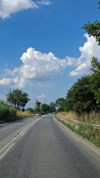 A perspective shot from a vehicle traveling on a two-lane asphalt road, bordered by dry grass and vegetation on the right, and green foliage and trees on the left. The sky is a vivid blue, dotted with fluffy white clouds. Several vehicles are visible on the road ahead, traveling in the same direction. The environment appears to be rural, with power lines and utility poles in the background. The scene is captured under daylight.