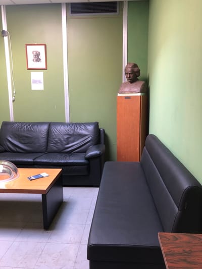 The image shows a room in a building located at 37.998 latitude and 23.82 longitude in Agia Paraskevi, Greece. There are two black leather couches, one against the wall and the other attached to the wall at a 90-degree angle. A wooden shelf is attached to the wall, and on it is a bust. There is a picture hanging on the wall next to the bust. Another picture is hanging on the wall to the left of the couch. A small, black table is attached to a larger wooden table against the wall next to the couch. An object is sitting on top of the wooden table. The room is painted light green. The floor is tiled. The wall is decorated with white horizontal trim. There are no people in the image.