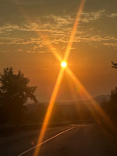 This is a photograph of a sunset over a rural landscape, likely taken from a road. The sun is positioned in the upper center of the frame, emitting bright rays of light that streak across the sky. The sky is filled with wispy clouds that are illuminated by the warm, orange hues of the sunset. In the foreground, a dark asphalt road curves away into the distance, marked by a white line. To the left of the road, a silhouette of a deciduous tree is visible. The background features rolling hills and distant mountains, softened by the atmospheric haze. The overall impression is one of a serene and picturesque natural scene at dusk. The image does not contain any people or specific urban elements of Beli Osam, Bulgaria.