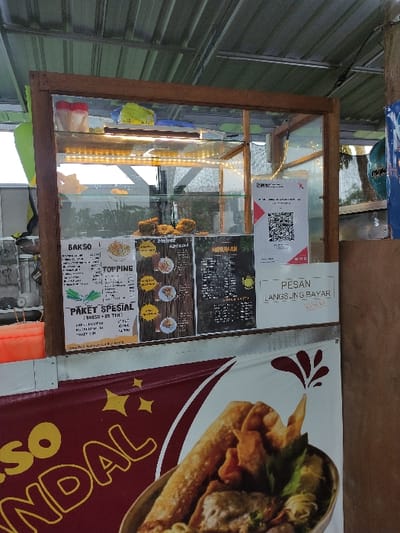 This image captures a vibrant street food stall, likely in Kecamatan Pandaan, Indonesia, during daytime. The central subject is a wooden-framed display case with glass panels, illuminated from within by a string of warm LED lights. Inside, various fried food items, possibly fritters or snacks, are showcased for sale.
To the right of the display case, a large banner with a maroon background and yellow text prominently displays "SO NDAL" (likely part of "Bakso Ndal"), alongside an appetizing graphic of a bowl of food or a wrap. Several laminated menus and signs are affixed to the back of the display case. One notable sign features a QR code for digital payments and the text "PESAN LANGSUNG BAYAR," meaning "Order, Pay Directly."
Other visible menu items include "BAKSO" (meatballs), "TOPPING," and "PAKET SPESIAL" (Special Package), with one option listed as "BRESO + ES TEH" (Breso + Ice Tea). The background on the left shows a corrugated metal wall or roof, characteristic of an outdoor or semi-permanent stall. Through the glass panels, distant trees and foliage are visible, indicating an open-air setting. No people are present, but the setup clearly indicates a ready-to-operate food vendor. The overall scene suggests a casual and accessible dining option.