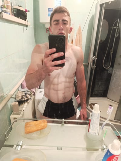 A man with defined abdominal muscles and prominent veins in his arm is taking a mirror selfie in a bathroom. He is wearing black and white shorts. A black smartphone is held in his right hand, obscuring his face partially. To his left, a white toilet is visible, and a radiator with towels draped over it. On the shelf in front of the mirror, there is a piece of orange food on a clear dish, a tube of toothpaste, a toothbrush, and a cotton swab. To the right of the man, a shower cabin with a sliding glass door is reflected in the mirror. The bathroom walls are tiled with light blue square tiles. The overall environment appears to be a personal bathroom, possibly in a residential setting within Fastiv, Ukraine, given the information provided in the prompt.